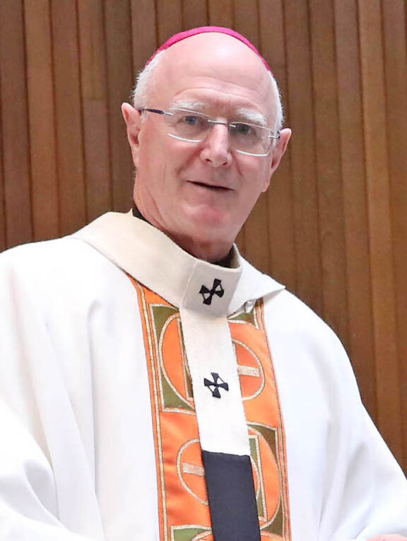 Picture of Archbishop Dermot Farrell in a white, orange, green and black robe wearing a pink skullcap smiling towards the camera in front of a brown wall.