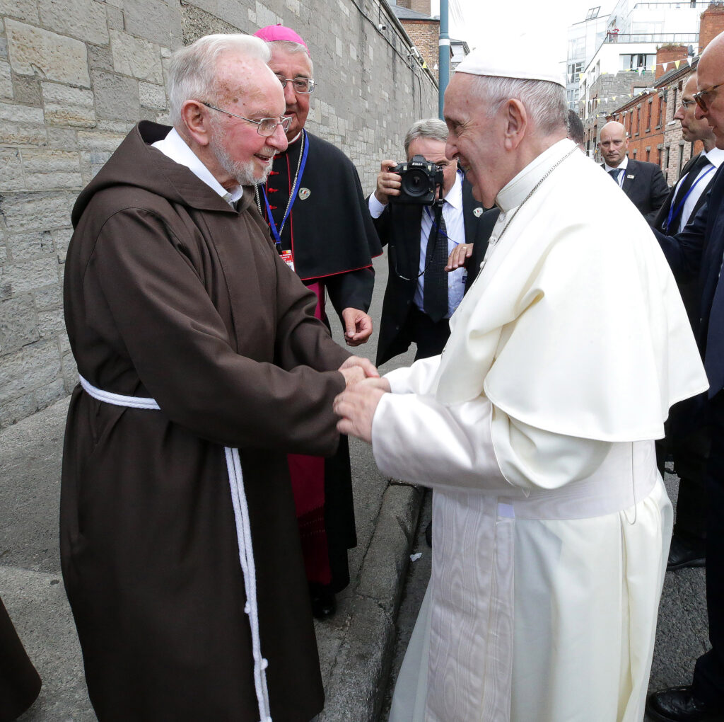 Pope Francis shakes hands with Br Kevin Crowley as he arrives at the Capuchin Day Centre on Bowe Street in Dublin. Archbishop Farrell delivers a statement on Wednesday 2nd of July 2025 extending his deepest sympathies to Brother Kevin's family and Capuchin Brothers on hearing about Brother Kevin's passing. In the photo we see Brother Kevin and Pope Francis in their different robes shaking hands. Behind them a man is taking a photo and other men and a priest are watching the handshake happen.