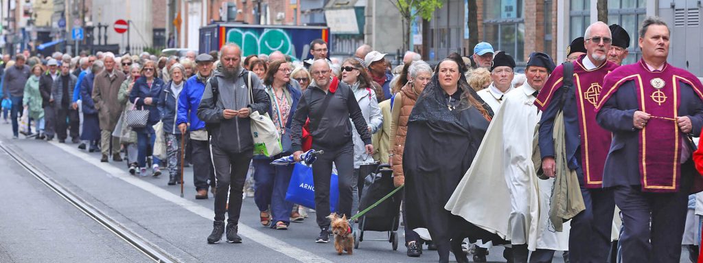 Graphic of a group of people walking on the street beside the Luas lines following two men in black and red suits. There is graffiti in the background. The Mass is to Commemorate the 100th Anniversary of the Death of Venerable Matt Talbot.