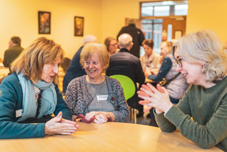 Graphic showing three women seated around a table with their hands together talking and smiling to each other. In the background there are yellow walls, two paintings on the walls a pair of brown fire doors and a table of other people talking to each other.