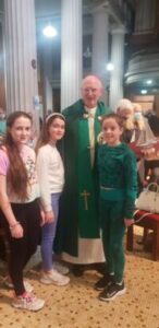 Picture of Archbishop Dermot Farrell wearing a green group standing with three girls to get their photo taken. All are smiling at the camera and the background is the inside of the church. This graphic is for the Opening Liturgy for the Diocesan Synodal Pathway.