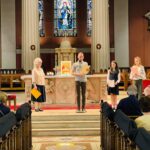 Graphic of a man in a blue shirt and black pants speaking to the congregation through a microphone from the altar.
