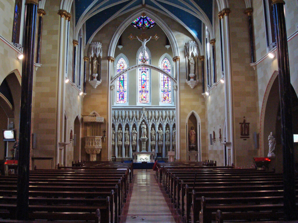 Solemnity of Christ the King – Homily of Archbishop Farrell in Blackrock. Graphic shows the inside of Blackrock Church with statues and stained glass windows in the centre background and benches for the congregation from the foreground to the background. There is a statue of Jesus towards the ceiling.