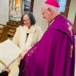 Graphic of a priest in a purple robe standing with a woman in a white jacket and scarf with a black top looking at a book on the stand of the altar.