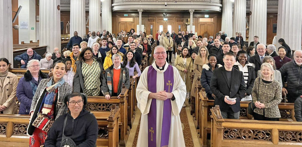 Graphic of the congregation standing to get their photo taken with Fr. Gareth Byrne who is wearing a purple and white robe over his black priest suit and white collar. The group are smiling at the camera.