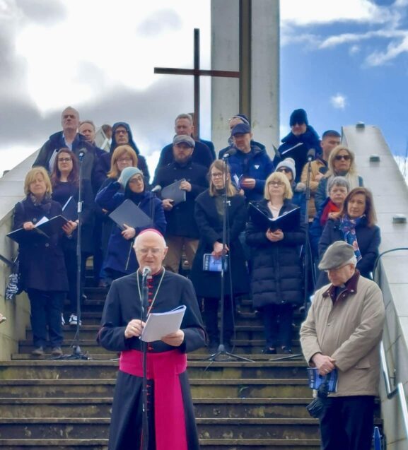 Graphic of The Way of the Cross Reflection of Archbishop Dermot Farrell which took place in Phoenix Park, Dublin on Good Friday, March 29, 2024. Here Archbishop Farrell is wearing a black and red priest suit and pink skullcap addressing the congregation through a microphone. Beside him is a man bowing his head in prayer and behind him is a choir with their songbooks open standing in front of a Holy Cross.