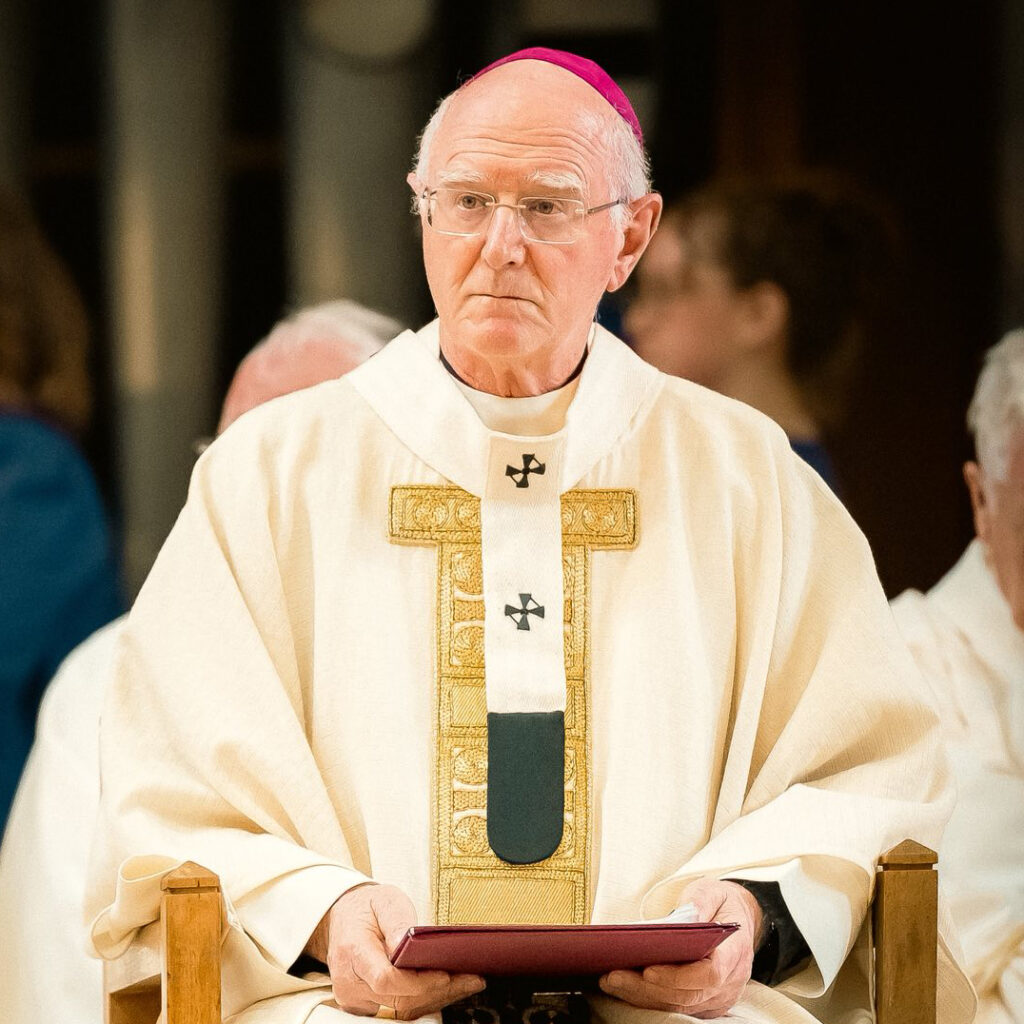 Graphic of Archbishop Dermot Farrell when he stated in public that it was time to address unprovoked attacks on the Indian community in Dublin. Here he is wearing a white and gold robe and a pink skullcap and is holding a red folder in his hands. In the background there are other members of the priesthood and people.