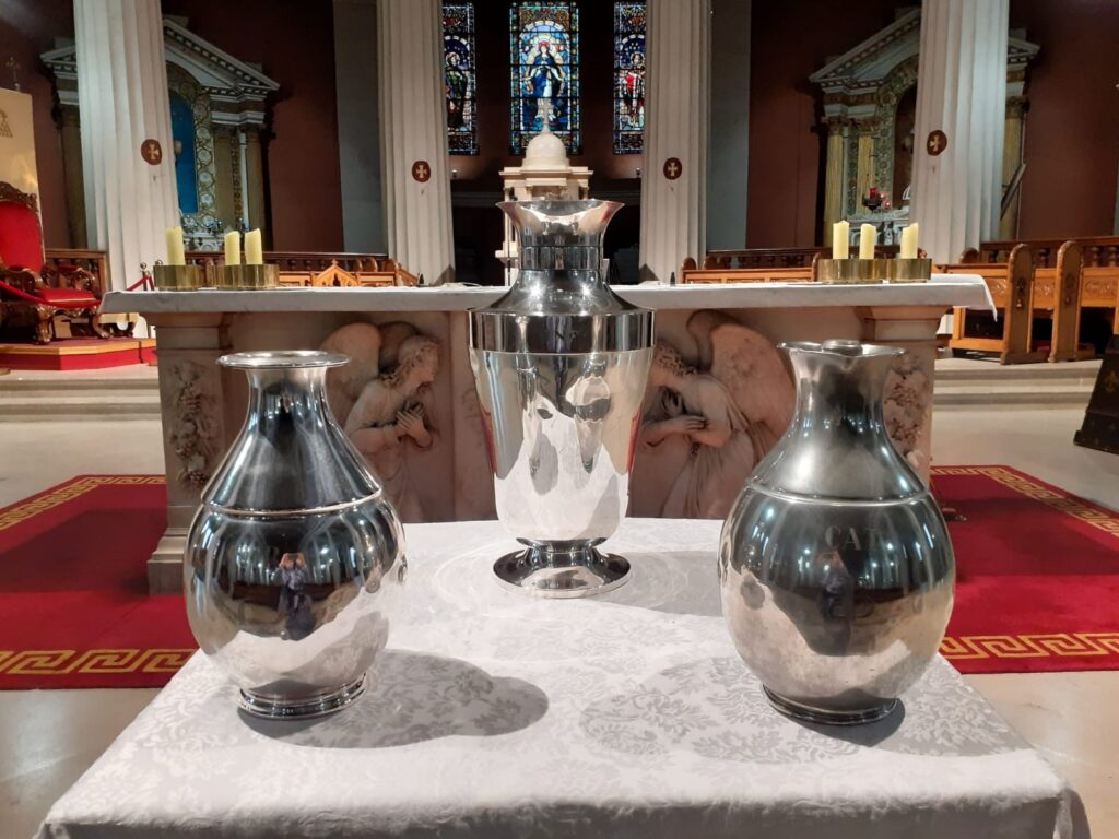 Christmas Mass 2023 – Homily of Archbishop Farrell. Graphic depicts three silver chalices on a table in front of the altar with a stained glass window and statues behind the table.