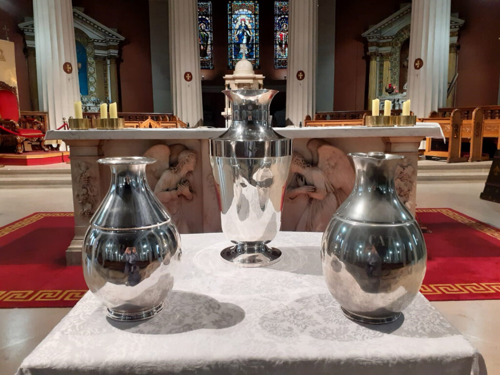 Graphic showing three chalices on a table at the altar which has carvings of angels and stained glass windows behind it.