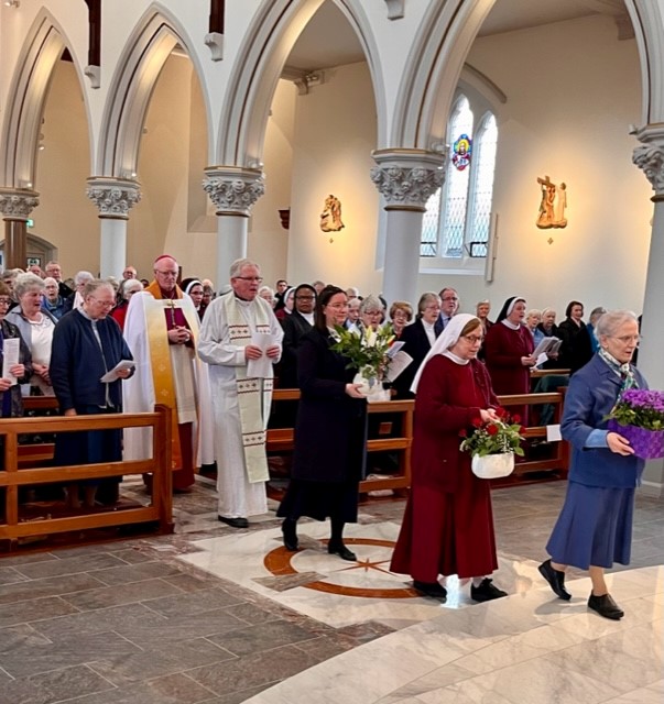 Day for Consecrated Life – Homily of Archbishop Farrell. Graphic shows three nuns bringing flowers to the altar and two archbishops behind them.