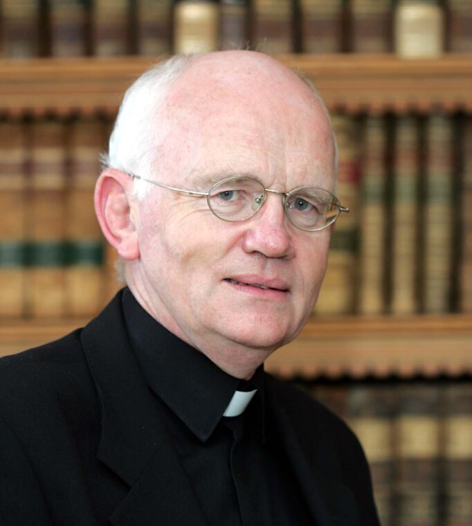 450th Anniversary of the Hospitaller Order of St John of God – Homily of Bishop Éamonn Walsh. Graphic depicts Bishop Éamonn Walsh smiling at the camera in a priest's suit. In the background there are bookshelves full of Bibles.