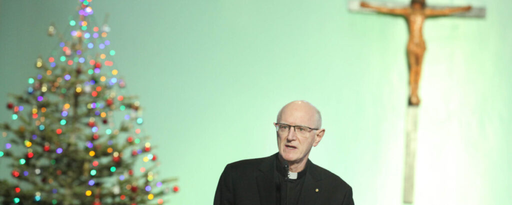 Graphic of Bishop Dermot Farrell's address on his appointment to Archbishop in the Church of Our Lady of Lourdes. He is wearing glasses and a black priest suit and is standing beside a Christmas tree and a cross with a statue of Jesus.