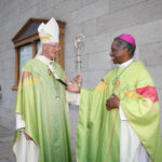 Graphic of Archbishop Farrell dressed in a green archbishop's gown with a headpiece talking to His Excellency Archbishop Jude Thaddeus Okolo, Apostolic Nuncio to Ireland wearing the same clothing but with a pink headpiece. They are in front of a white wall which holds the church notices.