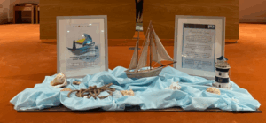 Synod display with a model sailboat, lighthouse, shells and framed Synod prayers on a blue fabric.