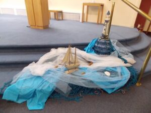A Parish Synod display set on the floor with a model sailboat and a tall striped lighthouse. White and blue fabric is draped to resemble waves, with seashells and netting scattered across the scene. The display is placed near the altar steps of the church.