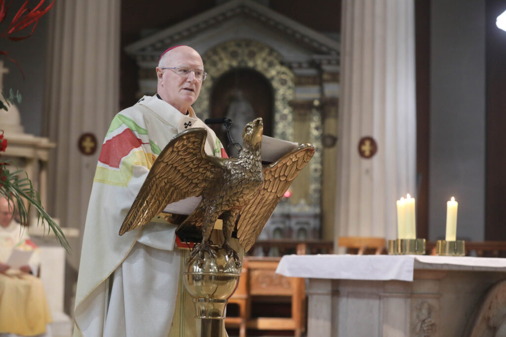 Graphic of Archbishop Farrell addressing the congregation in a white, red, green and yellow robe and pink skullcap from the altar's stand in the shape of a dove.