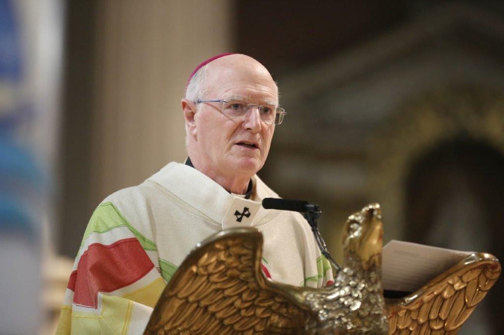 Graphic of Archbishop Farrell addressing the congregation in a white, red, green and yellow robe and pink skullcap from the altar's stand in the shape of a dove.