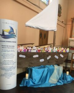 A parish Synod display with a large wooden boat holding colourful cut out figures of people, each labelled with words like 'Knowledge', 'Courage', 'Love', 'Care' and 'Wisdom'. A white sail rises above the boat with blue fabric and candles arranged below to represent water. To the left stands a banner with the Synod logo and prayer.