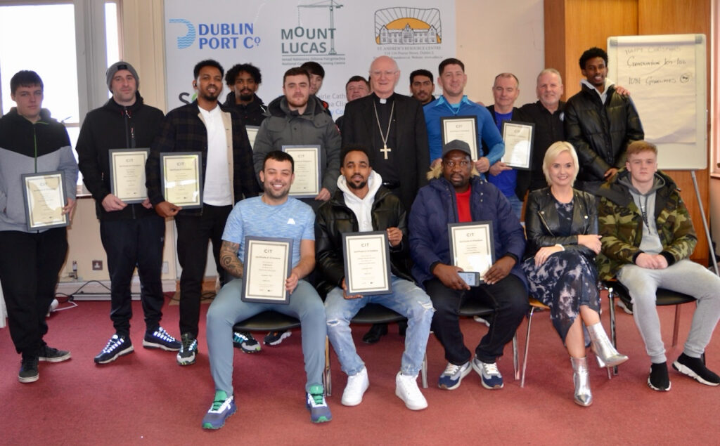 Graphic of the presentation of certificates at the Construction Skills Programme in Dublin Port which Archbishop Farrell remarked at. The people are all smiling at the camera many with their certificates and Archbishop Farrell is in the centre in his black priest suit and white collar with a gold cross around his neck.