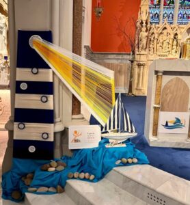 A parish Synod display inside a church with a model sailboat on blue fabric surrounded by stones. To the left, a dark panel with circular decorations has yellow and orange threads stretched diagonally like rays of light. A Synod logo poster is placed in front, with a carved altar and stained glass windows visible in the background.