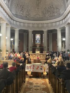 Graphic shows the congregation in a church sitting on benches with some people sitting on the altar listening to a man speak from the stand on the altar for the Opening Liturgy for the Diocesan Synodal Pathway.
