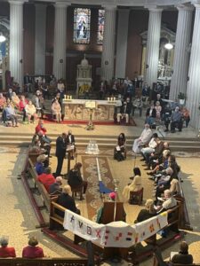 Graphic shows a birds-eye view of a group of people gathered on benches and seated on the altar listening to a man in a black suit speak from the stand in a church for the Opening Liturgy for the Diocesan Synodal Pathway.
