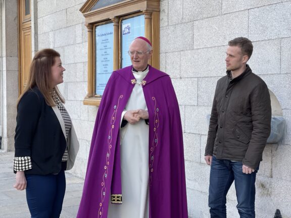 Graphic of the Lay Ministry Collection - 14th/15th May. Graphic shows a priest in a white and purple robe talking to a woman and a man in front of the billboard of the Church beside the Church doors.