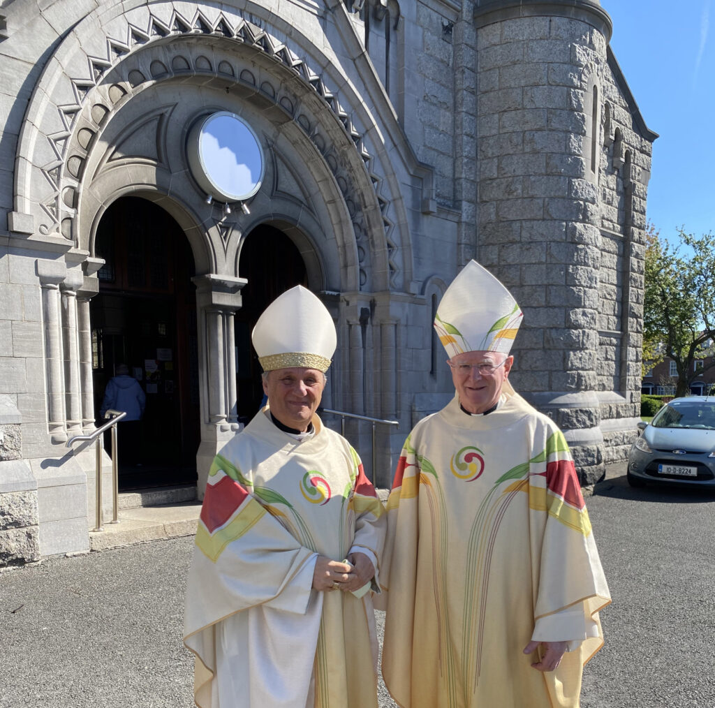 Picture of Cardinal Mario Grech and Archbishop Dermot Farrell standing beside each other in white, green, yellow and red robes with headpieces outside of a church with a car in the background. The two men are smiling at the camera.