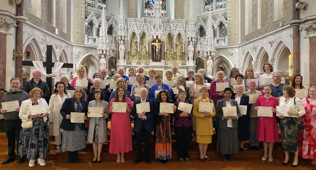 Graphic of the 45 people who completed the year-long Certificate course in Catechesis getting their photo taken holding their certificates with Archbishop Dermot Farrell in the background in his green, white, yellow and red robe in front of the altar. They are all smiling at the camera.
