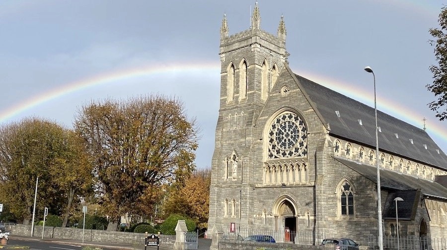 Graphic of the Church of the Sacred Heart in Donnybrook with two cars outside the church and a rainbow and trees to the left of the image. There is a clear blue sky behind the church and a black sign in front of the church.