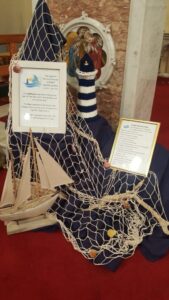 A Parish Synod display with a small wooden sailboat model and a black-and-white striped lighthouse. A fishing net with shells is draped across dark blue fabric, with two framed explanation cards describing the Synod logo and prayer. In the background is a religious painting on the wall.