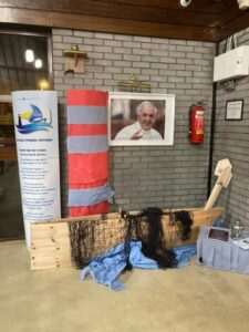 A parish display against a brick wall featuring a wooden boat frame with fishing net and blue fabric draped to resemble water. Beside it stands a red and grey striped buoy model and a banner with the Synod logo and prayer. Above the display is a photograph of Pope Francis, with a fire extinguisher mounted on the wall nearby.