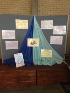 A parish display with blue and teal fabric draped to form the shape of a sail on a grey backdrop. The centre features a Synod logo with a boat, surrounded by handwritten posters and reflections pinned to the background. Additional posters are placed on stands in front of the display.
