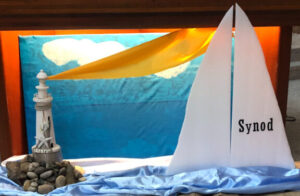 A church display featuring a lighthouse model on rocks beside a white sail with the word 'Synod' printed on it. The background is a colourful mural with blue sea, white clouds, and a golden sail like shape, while and blue fabric is draped below to resemble water.