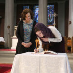 Graphic of two women at the altar with one woman signing a book and the other woman looking at the woman signing the book. There is stained glass windows and columns in the background. They are becoming Christians.
