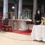 Graphic of two women at the altar wearing facemasks with one woman hunched over. The background is the altar with a red carpet and a red and brown chair. They are becoming Christians.