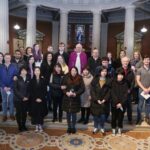 Graphic of a group of people standing during the Rite of Election 2022 with the priest. Their hands are together when they stand and they are looking towards the camera with a stained glass window of Mother Mary in the background amongst white pillars.