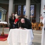 Graphic of a man signing the book to become Christian with a woman standing beside him holding a child. The priest can be seen to the right in a white robe.