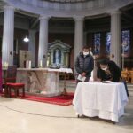Graphic of two people at the altar with one person signing the book and the other person looking at it. The background is the altar with white columns stained glass windows and a red carpet. They are becoming Christians.