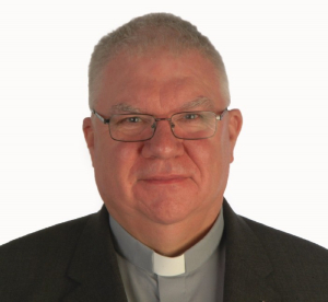 Picture of Mgr. Dan O’Connor who is the Episcopal Vicar for Education. Here Dan is pictured smiling at the camera, wearing glasses and a black jacket with a grey shirt and white collar and he has a white background.