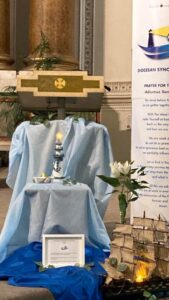 A church display with a lectern in the back and a decorative arrangement in front covered in blue fabric. This setup includes a lit candle on a stand with an anchor symbol, a framed picture at the bottom, a vase with white lillies, and a model ship on the right side. A banner in the background reads 'Diocesan Synod' with a logo of a boat and text beginning with 'prayer for Synod'.