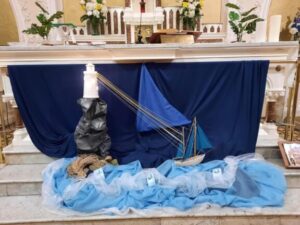 Synod display in a parish of the Archdiocese of Dublin featuring a church altar decorated with blue fabric, a model sailboat, a lighthouse with light, fishing net, and symbolic water effects.