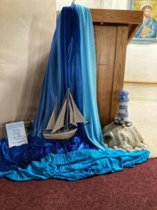 Synod display in a parish of the Archdiocese of Dublin featuring a wooden lectern draped with blue fabric, a model sailboat, a lighthouse decoration, and a printed prayer on display.