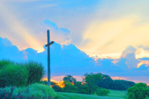Graphic shows a cross on a hill surrounded by greenery with a sunrise in the background with large white fluffy clouds.