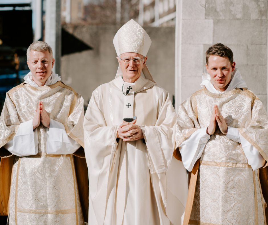 Graphic of Archbishop Dermot Farrell standing in the middle of the photo in a white robe and a headpiece with his hands clasped and two men to either side of him in white robes. Father Farrell is ordaining both of these men to the Diaconate. The background is a white wall and white stairs.