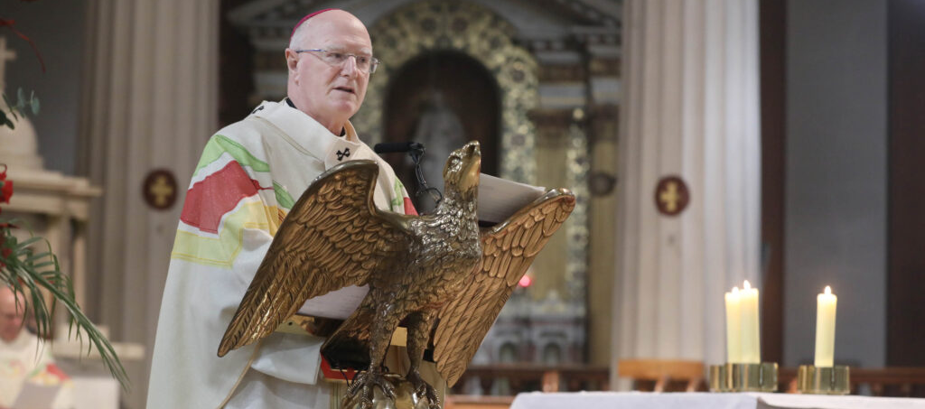 Graphic of Archbishop Farrell reading to the congregation from the altar stand in the shape of a dove. He is wearing a white robe with green red and yellow stripes and a red hat. In the background there is a marble statue and two Holy Crosses on white columns. There are two candles to the right of the graphic.