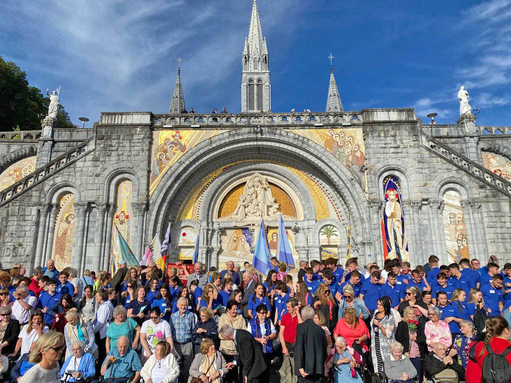 The congregation that attended the Homily of Fr Gareth Byrne at the Grotto of Lourdes for the Blessed Mary's birthday on the 8th of September. They are congregated in front of a marble statue which contains the Blessed Mary and two other figures from the Bible one of which is the Child Jesus. There are paintings of the apostles depicted around the statue. The Church spire can be seen in the background in the clear blue sky. On either side of the steps there are two other statues. There are flags on the ground behind the congregation.