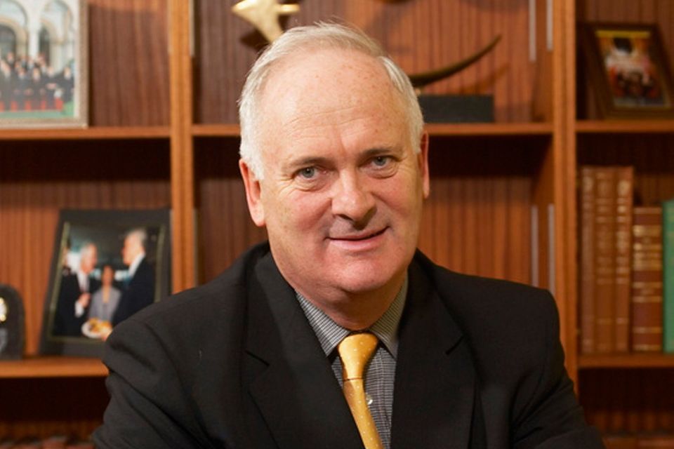 Picture of the deceased former Taoiseach John Bruton smiling towards the camera in a black suit with a blue and white shirt and yellow tie who is sat in front of bookshelves.