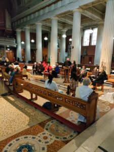 Graphic for the Opening Liturgy for the Diocesan Synodal Pathway. Graphic shows a group of people sitting on benches in a church with two people speaking to them from a stand in the middle of the room.