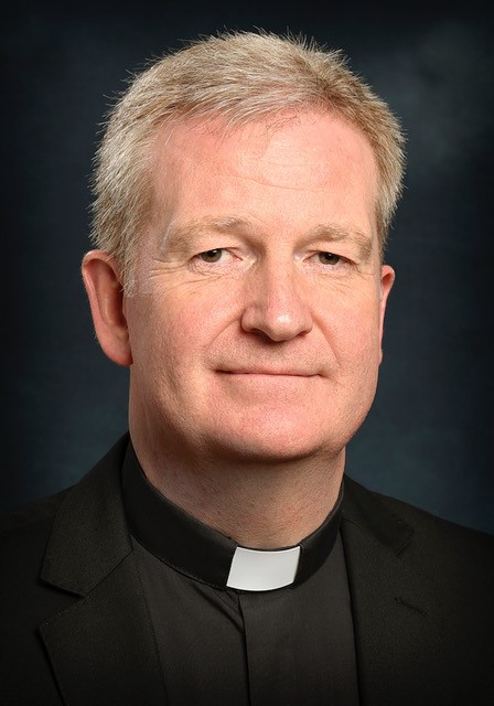 Picture of Paul Dempsey on his appointment from Bishop to Auxiliary Bishop of Dublin. He is pictured wearing a black priest suit and white collar and has a dark background. He is smiling at the camera.